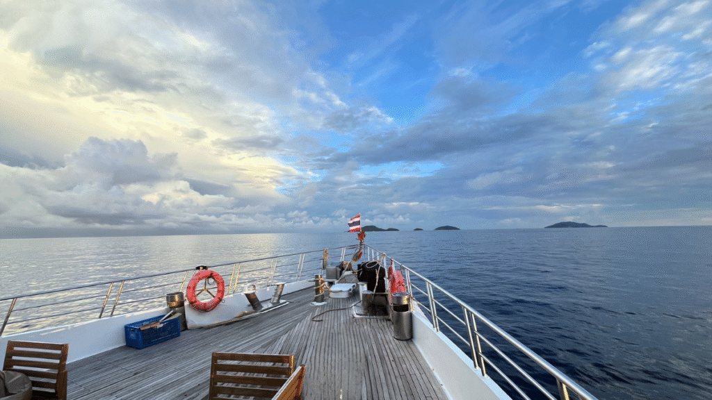 View from the wooden sundeck of a liveaboard boat, looking out at the open ocean and distant islands at dusk.