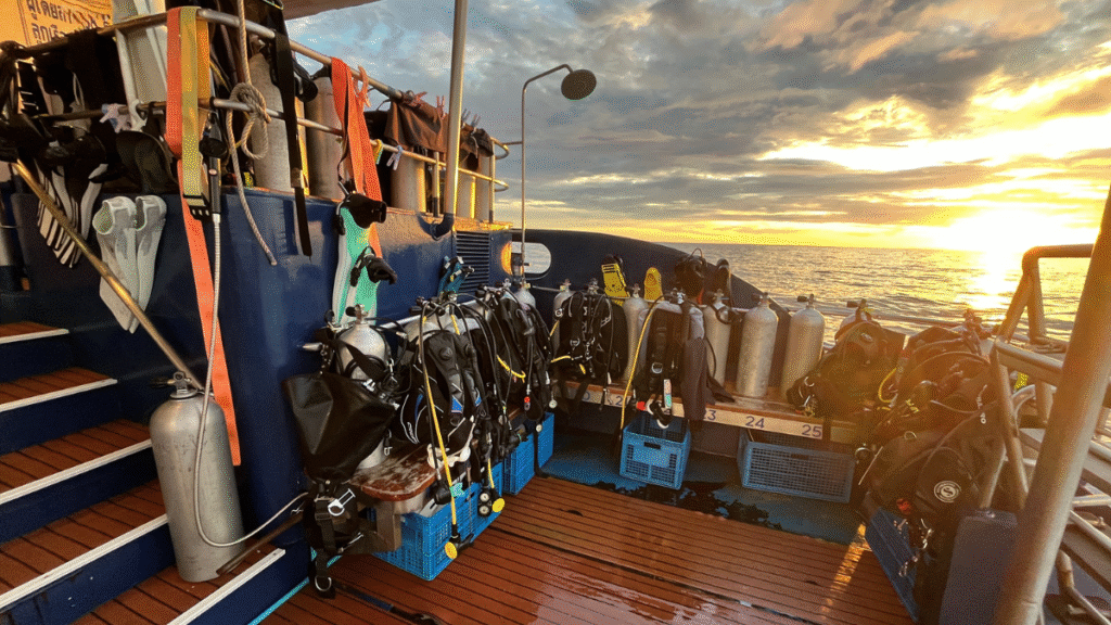 The dive deck of a Thailand liveaboard at sunset, fully equipped with scuba tanks and BCDs ready for a dive.