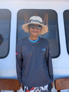 Bank, the MV Raga dinghy driver, wearing a wide-brimmed sun hat and sunglasses, smiling on the deck.