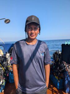 Portrait of Dan, a lively deck hand from Nakornpanom, standing on the dive boat deck wearing a navy staff shirt and black cap, smiling.