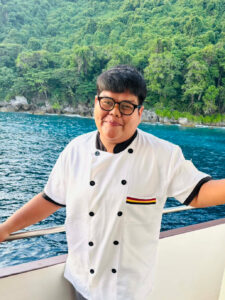 Fia, the head cook of MV Raga, smiling in his chef uniform with the ocean in the background.