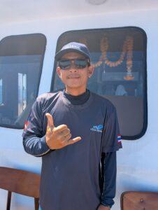 Kang, an experienced dinghy driver from Trang, giving a shaka hand sign while wearing sunglasses and a cap on the boat deck.