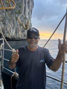 Portrait of Saeri, the boat engineer, wearing sunglasses and a cap, giving a confident thumbs-up on the deck with limestone cliffs in the background.